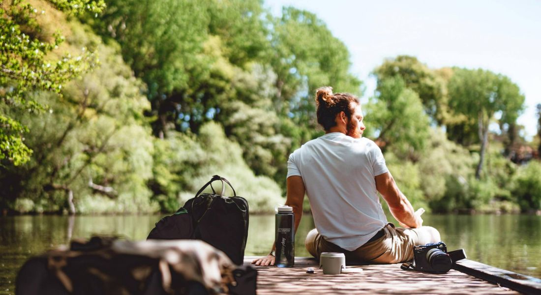 A person sits on a wooden dock by a lake, surrounded by trees, with bags and a thermos nearby on a sunny day.