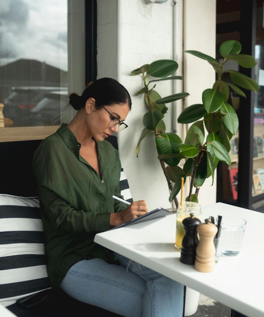 Woman in glasses writing on a tablet at an outdoor café table with a drink and plant nearby.