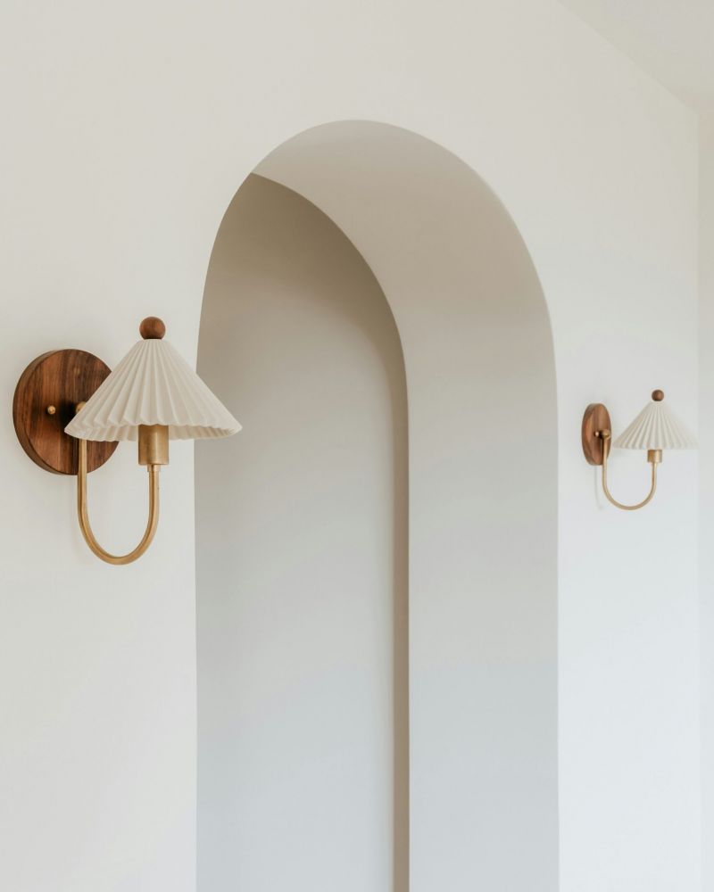 Minimalist wall lamps with pleated shades flank a white, arched doorway in a modern interior.