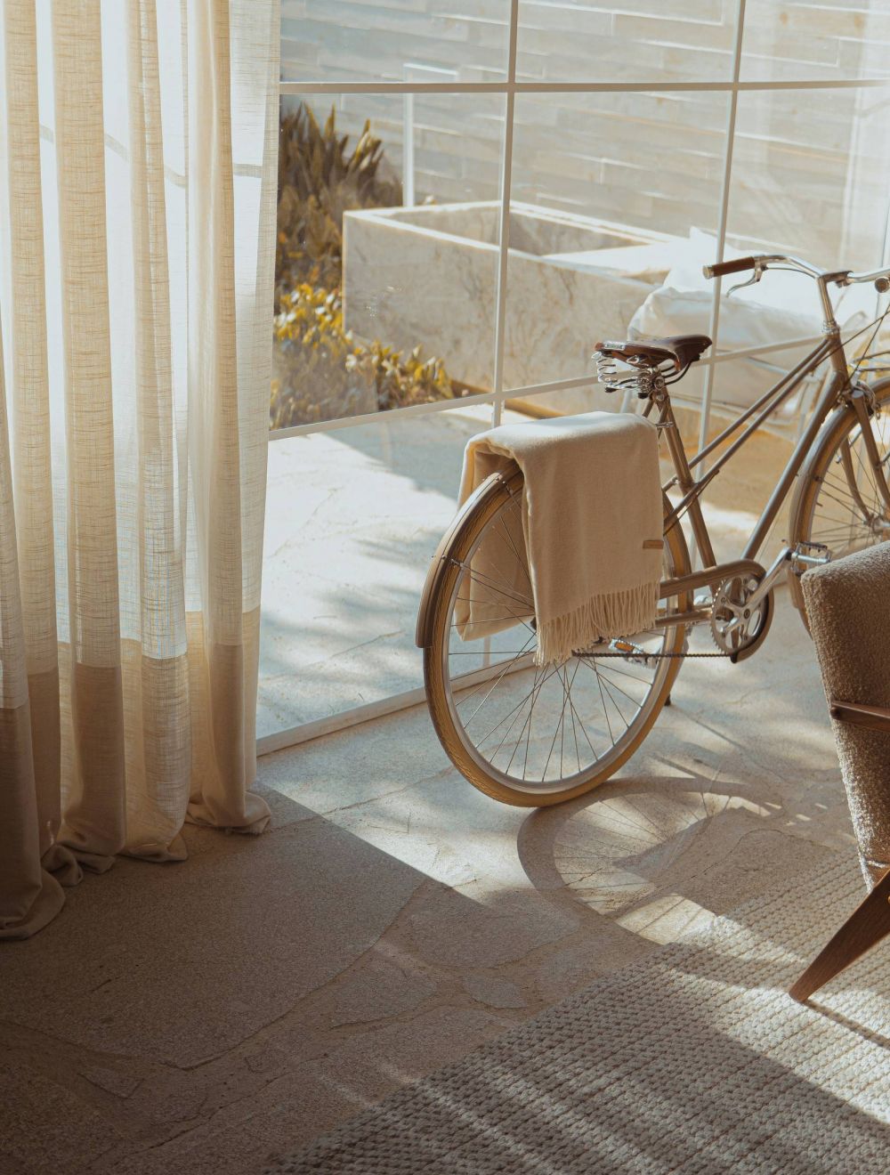 A bicycle with a beige blanket is parked indoors by large sunlit windows with sheer curtains.