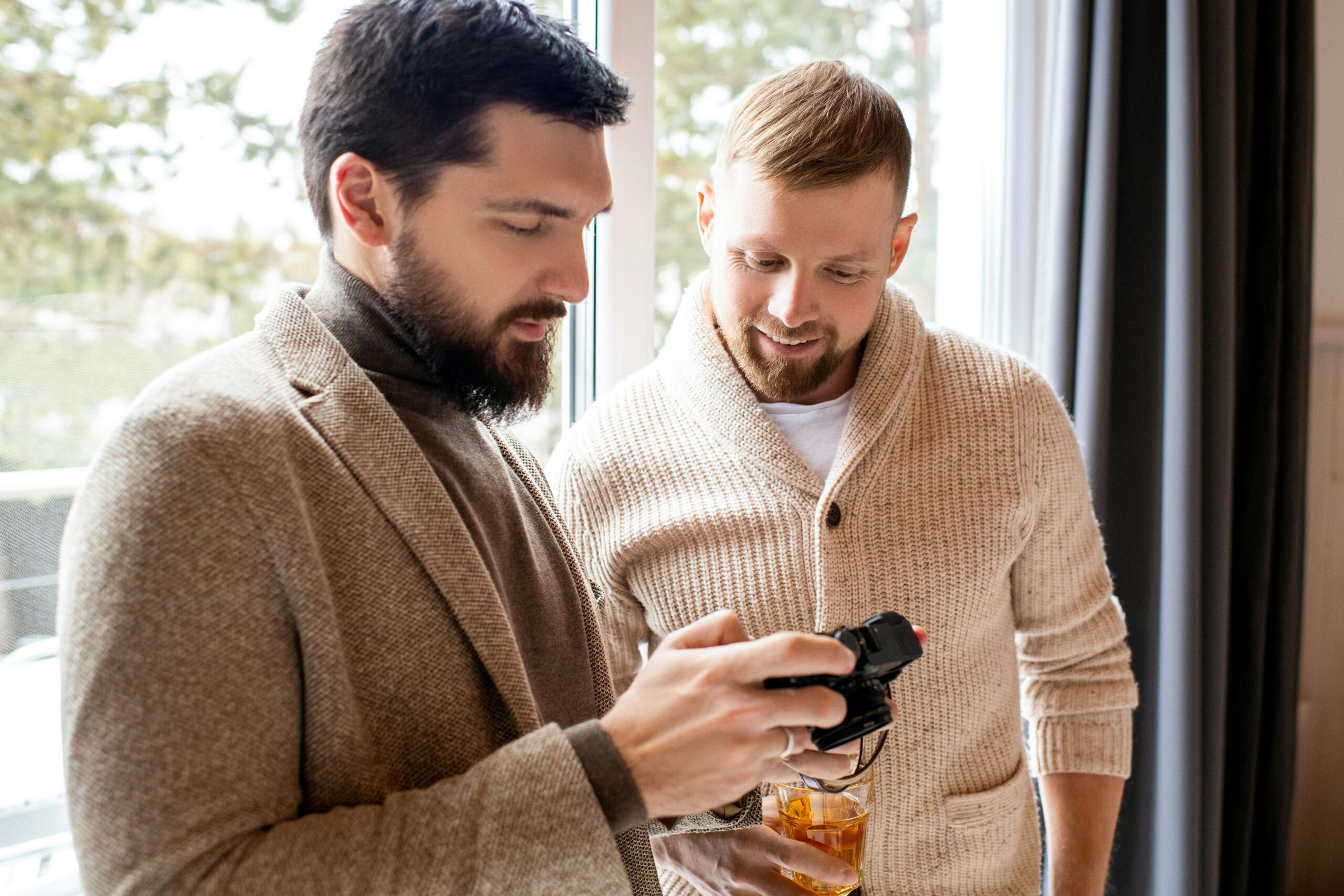 Two men stand by a window. One holds a camera and shows the screen to the other, who holds a drink and smiles.