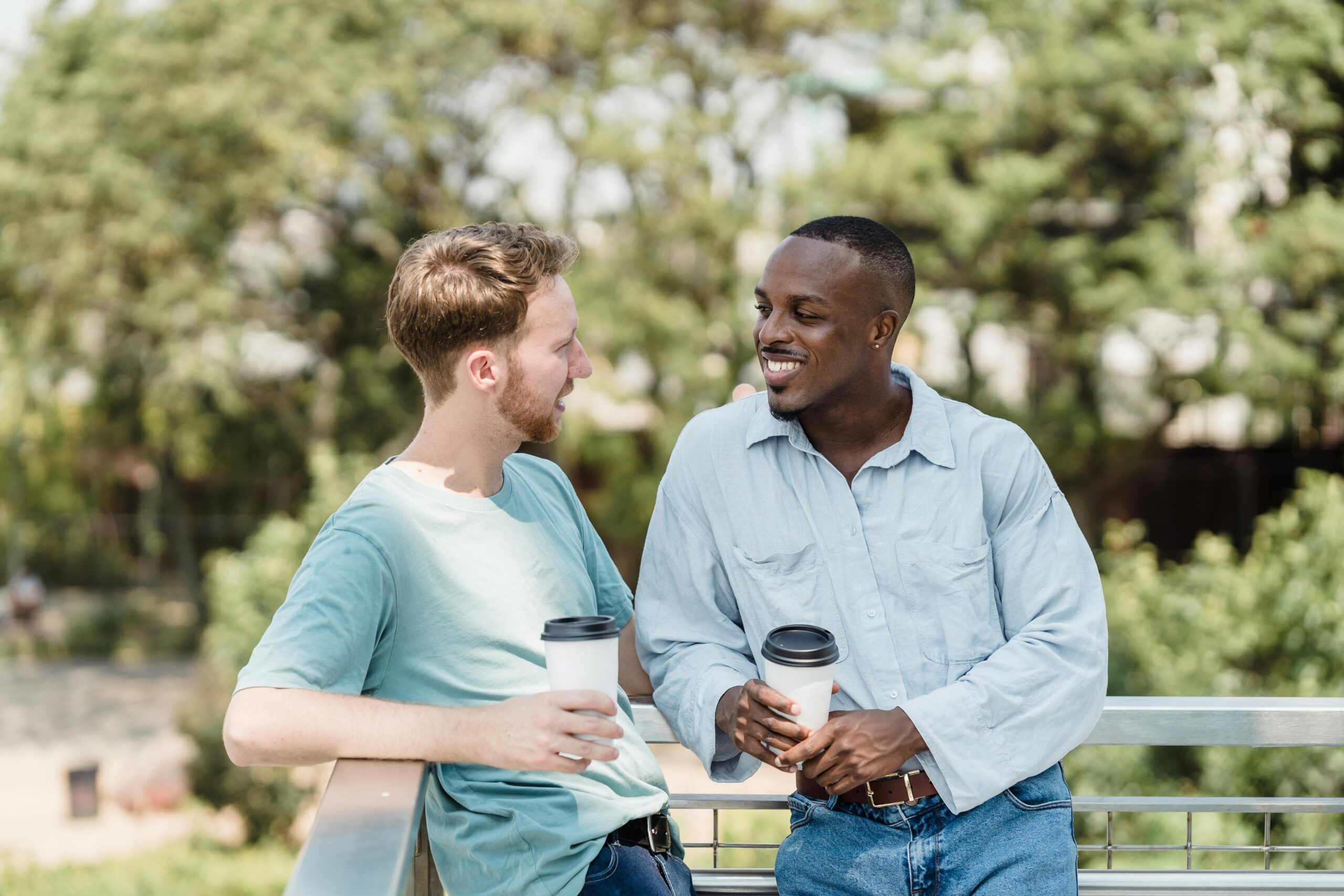 Two men stand outside, smiling and talking while holding coffee cups, with greenery in the background.