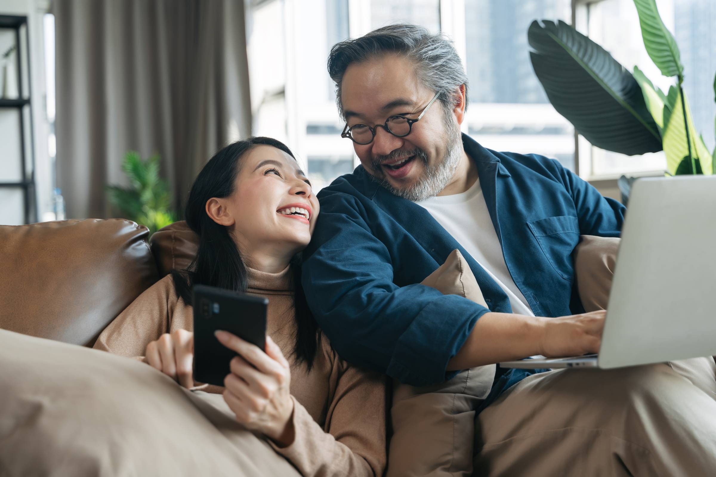 A smiling couple sits on a couch; one holds a phone, the other uses a laptop. They look at each other and laugh.