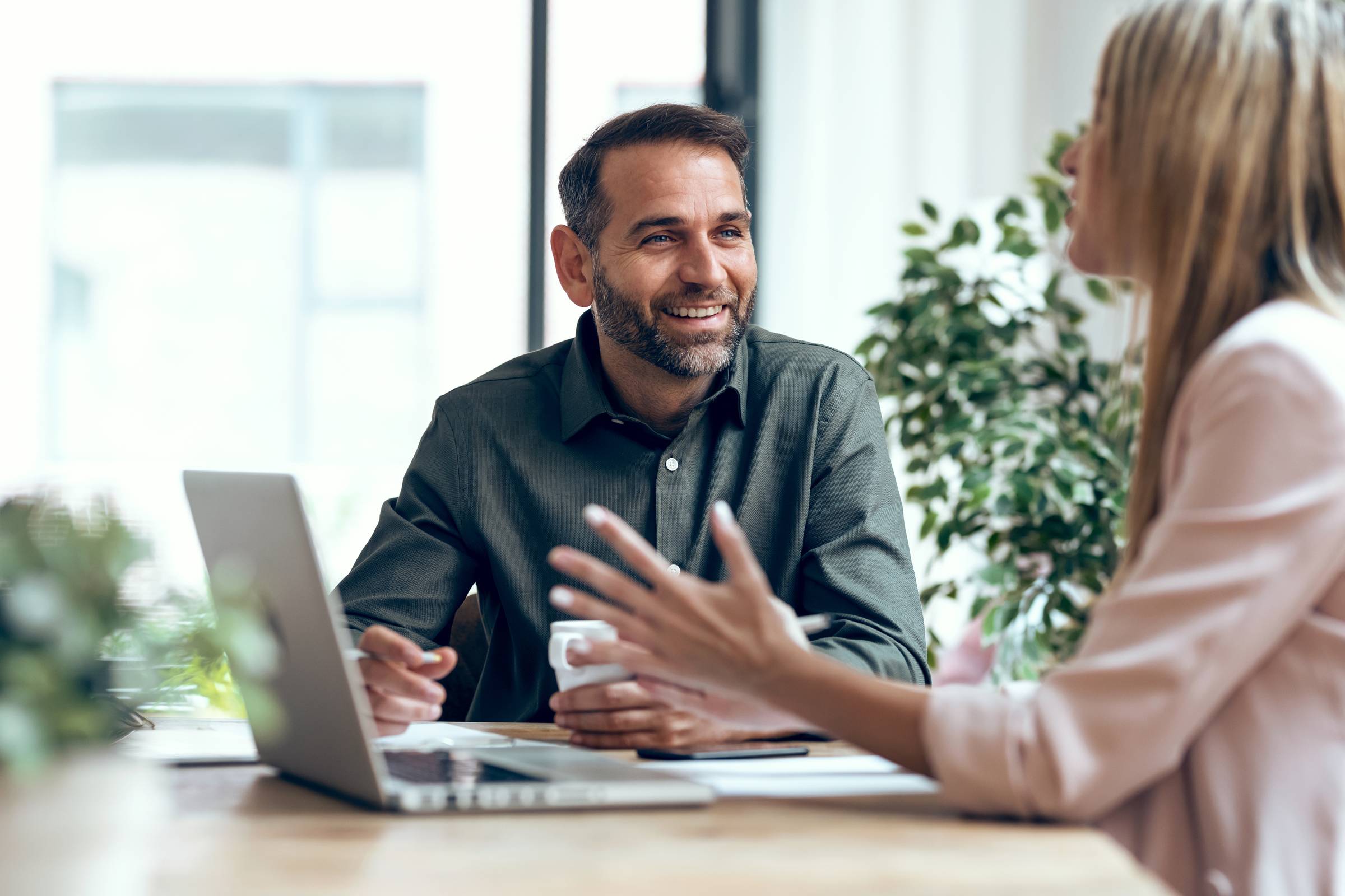 A man and a woman have a discussion at a desk with a laptop and paperwork, smiling in a bright office.