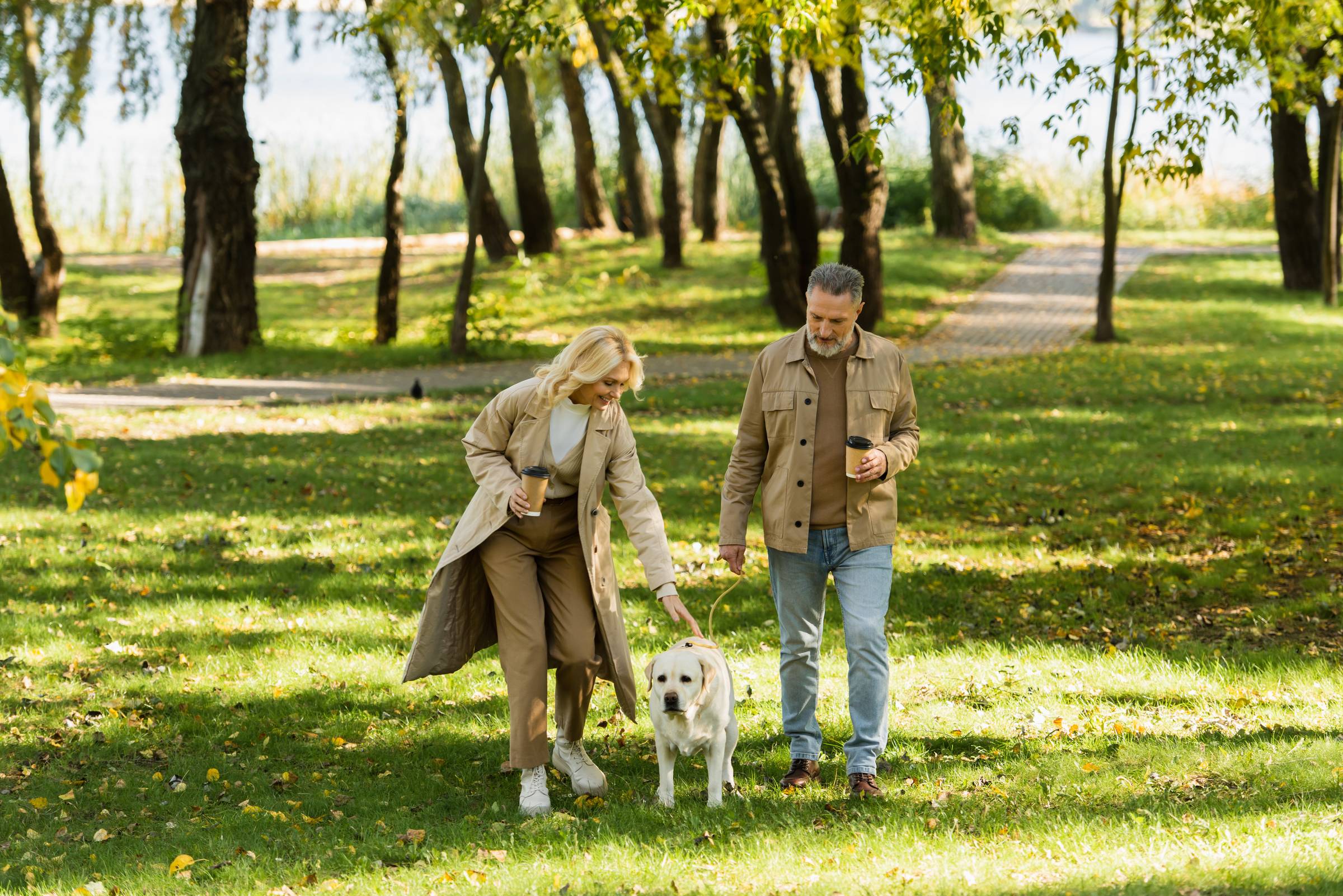 Older couple walking a yellow dog together in a sunny park, both holding coffee cups, trees and grass in the background.