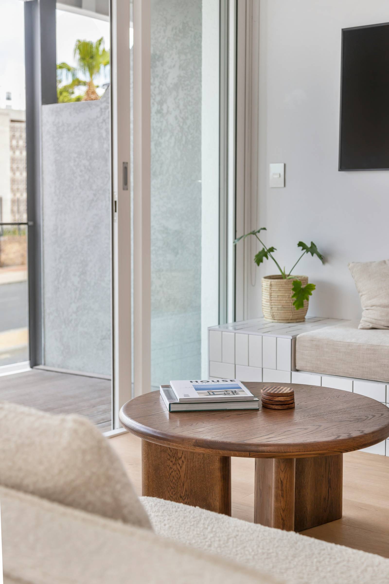 Round wooden coffee table with books, beige seating, potted plant, and large window in a modern living room.