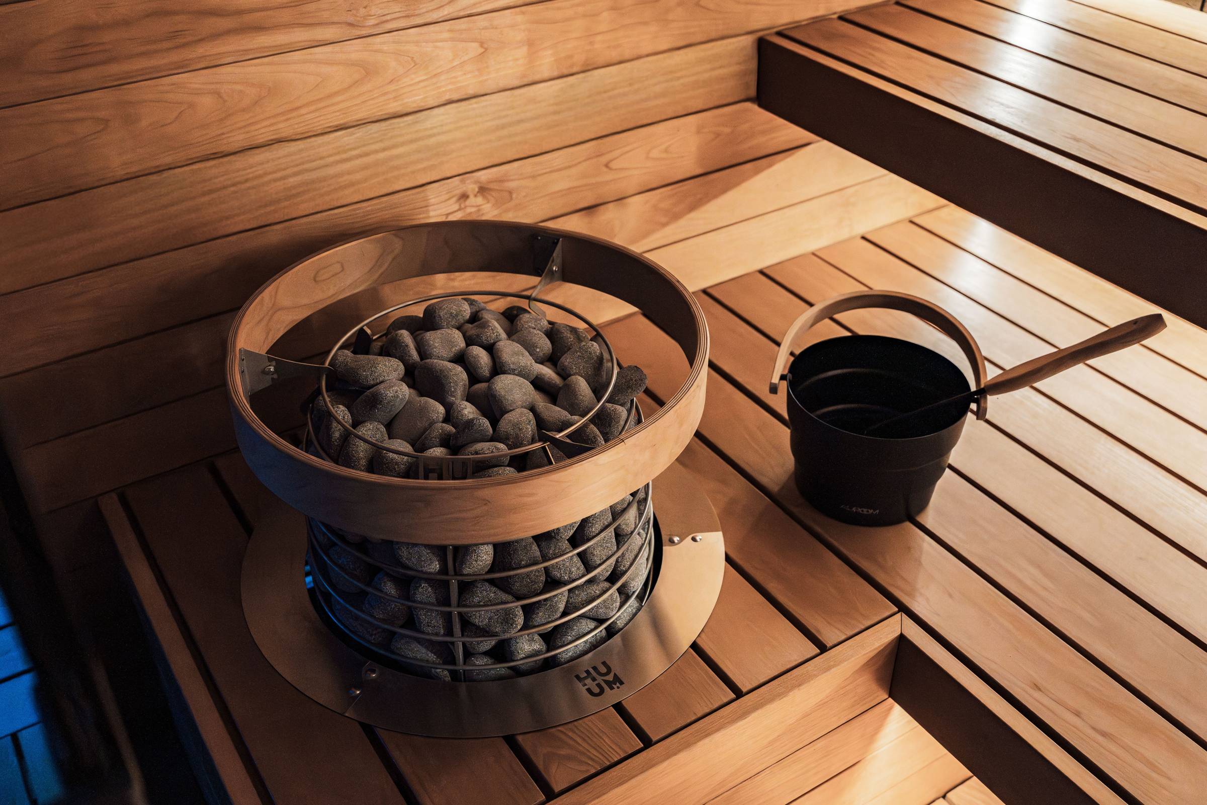 Modern sauna with a wooden bench, a round heater filled with stones, and a black bucket with a ladle on the wooden floor.