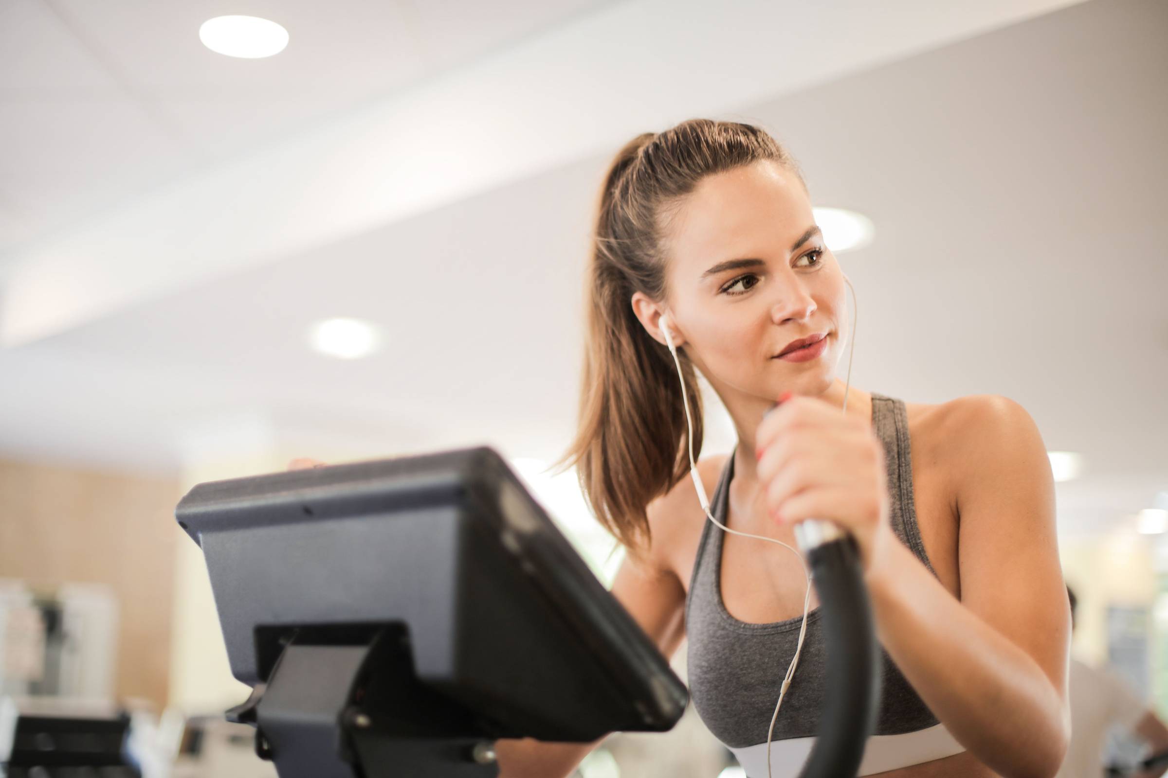 Woman in athletic wear using an exercise machine at the gym, listening to music with earphones, looking to the side.