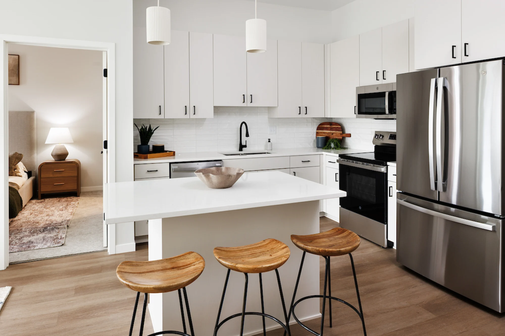 Modern kitchen with white cabinets, stainless steel appliances, island with three wooden stools, and pendant lights; bedroom visible.