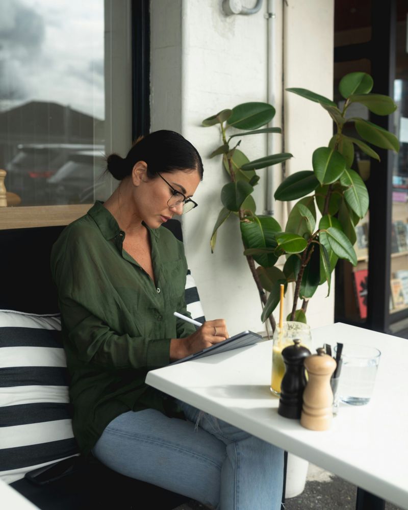 Woman in glasses writing on a tablet at an outdoor café table with a drink and a plant nearby.