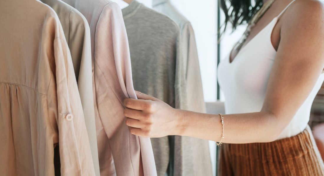 A woman in a white tank top browses light-colored clothes on hangers, touching a pink jacket.