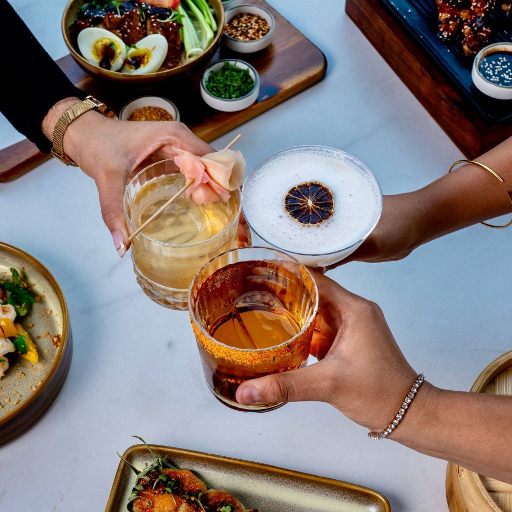 Three hands clinking cocktails over a table set with assorted food, including eggs and greens, on a white surface.