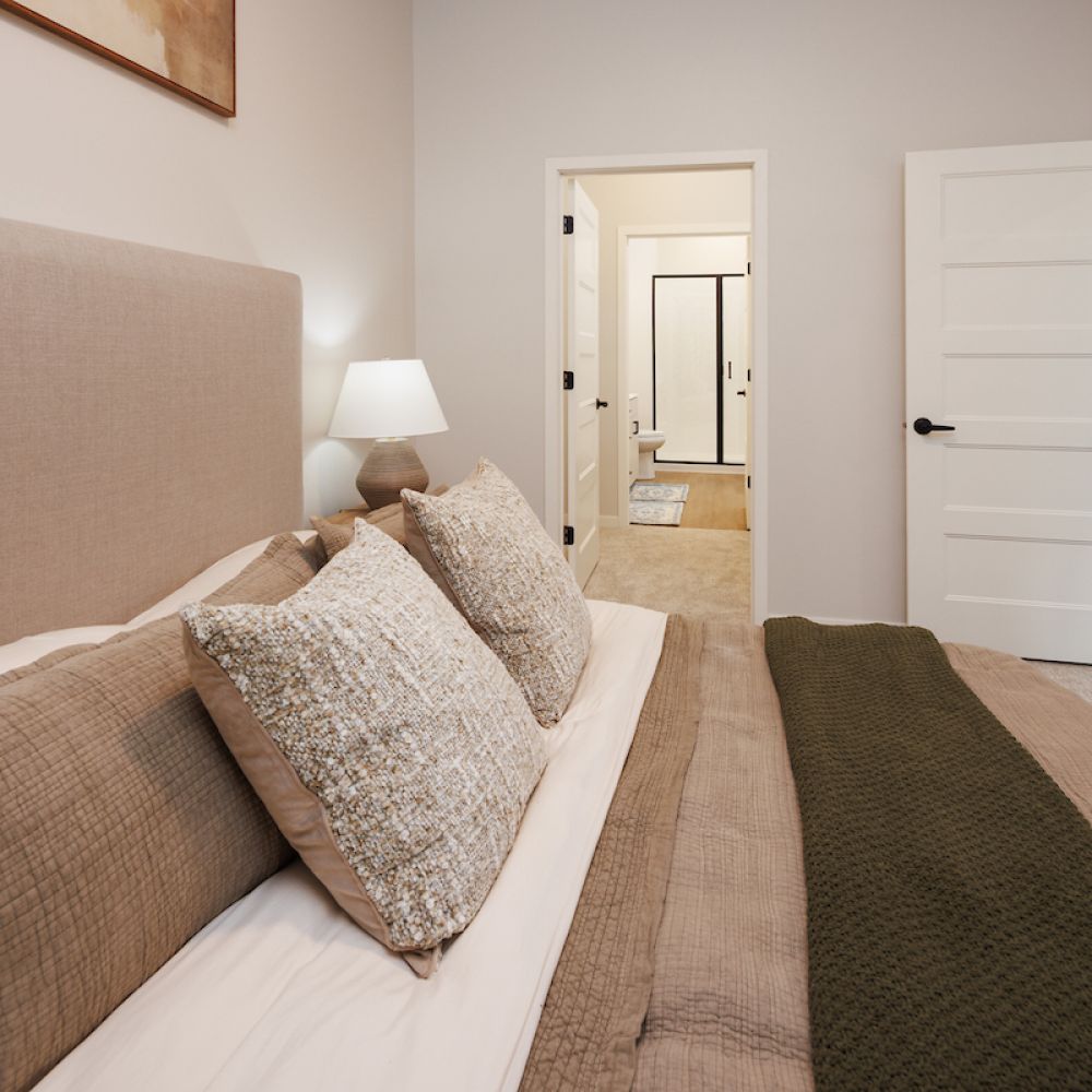 Bedroom with neutral bedding, lamp on nightstand, view of ensuite bathroom, and kitchen visible through open door.