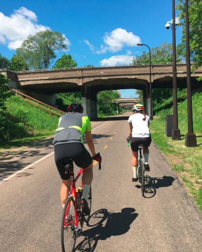Two cyclists ride on a sunny paved path under a bridge, surrounded by green trees and grass.