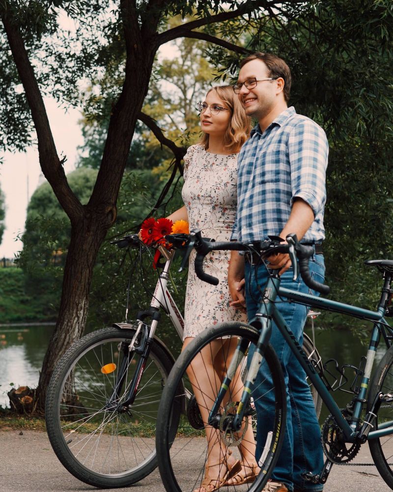 A smiling couple stands with their bicycles near a lake, surrounded by trees, enjoying a peaceful day outdoors.